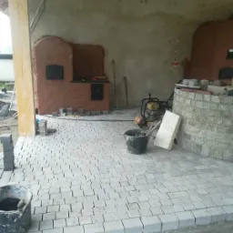 Niedokończony taras z szarej kostki granitowej, widoczne dwa piece chlebowe z cegły, wiadro z zaprawą, ubijarka wibracyjna i stos kostki do wykończenia nawierzchni.