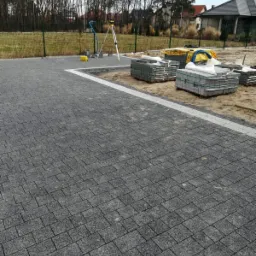 Ułożona, szara kostka brukowa z białym obramowaniem, widoczne palety z kostką, niwelator laserowy i ogrodzenie w tle.