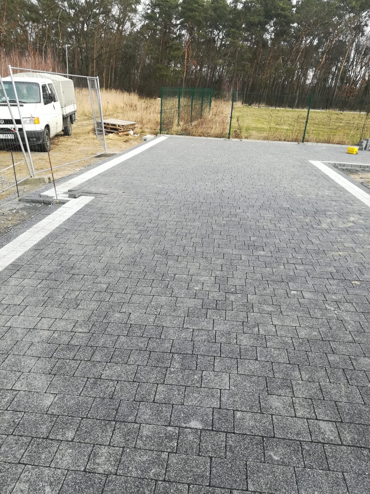 Ułożona szara kostka brukowa na podjeździe z białymi obrzeżami, widoczny samochód dostawczy i ogrodzenie w tle, częściowo ukończone prace wykończeniowe.