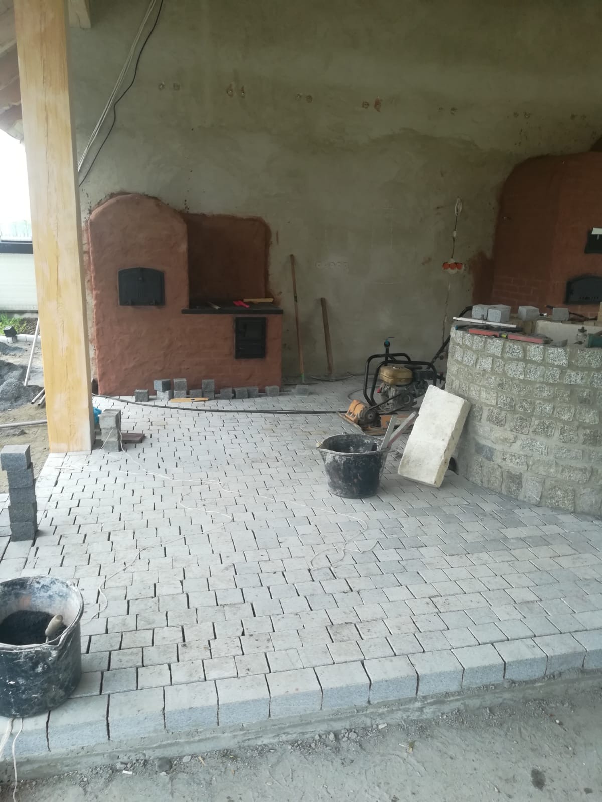 Niedokończony taras z szarej kostki granitowej, widoczne dwa piece chlebowe z cegły, wiadro z zaprawą, ubijarka wibracyjna i stos kostki do wykończenia nawierzchni.