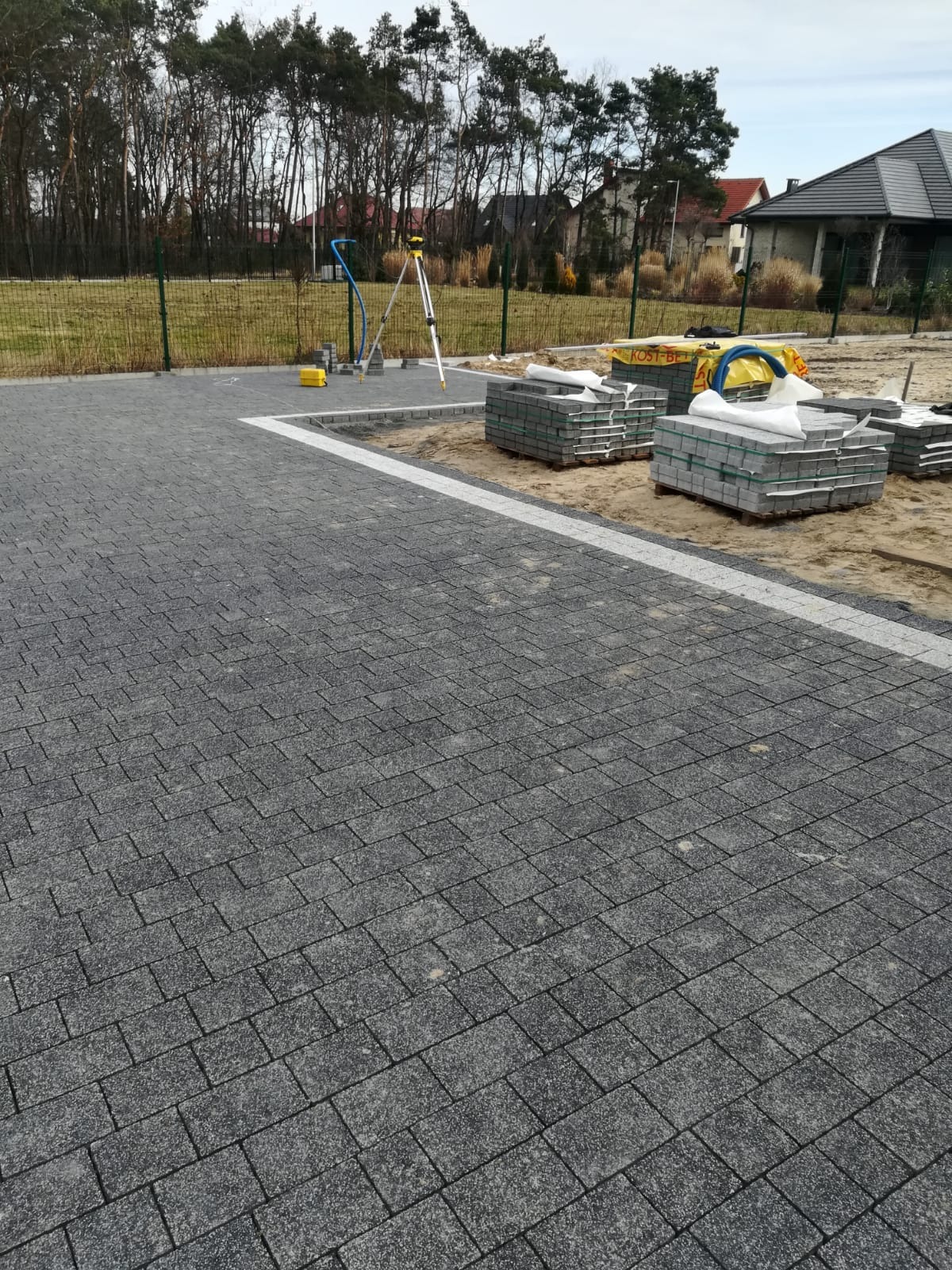 Ułożona, szara kostka brukowa z białym obramowaniem, widoczne palety z kostką, niwelator laserowy i ogrodzenie w tle.