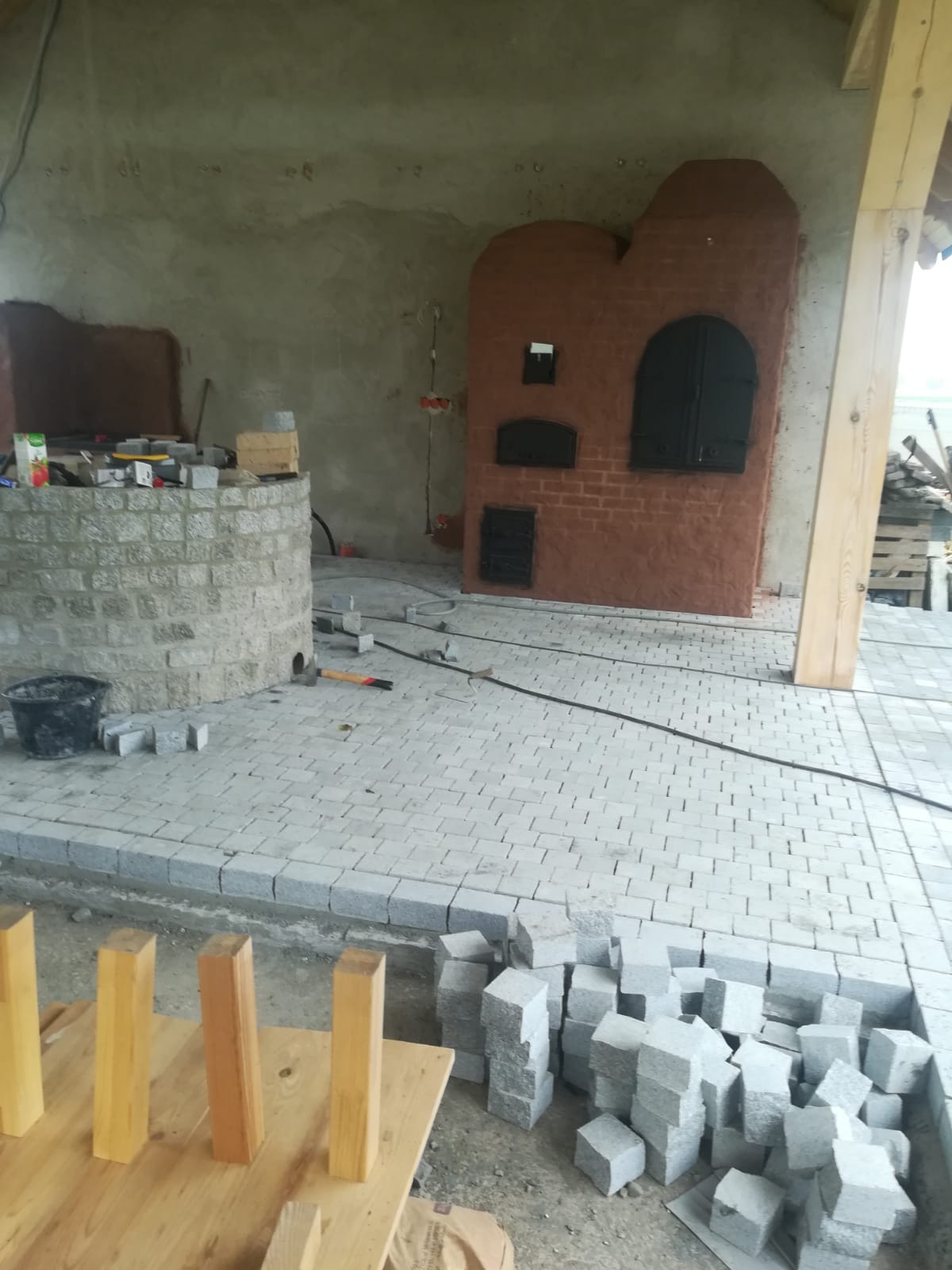 Układanie jasnoszarej kostki brukowej wokół murowanego pieca w Prudniku, widoczne narzędzia i materiały budowlane, częściowo ukończona powierzchnia.