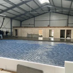 Montaż ogrzewania podłogowego w hali przemysłowej, widoczne niebieskie rury na izolacji oraz wylewka w tle, pracownicy zacierają wylewkę przy ścianie.