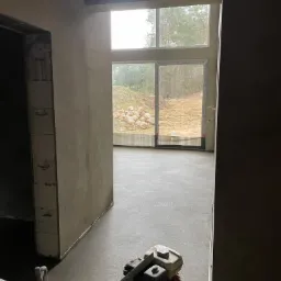 Świeżo wylana, szara posadzka w surowym wnętrzu, widoczna maszyna do zacierania betonu, duże okno z widokiem na ogród z kamieniami.