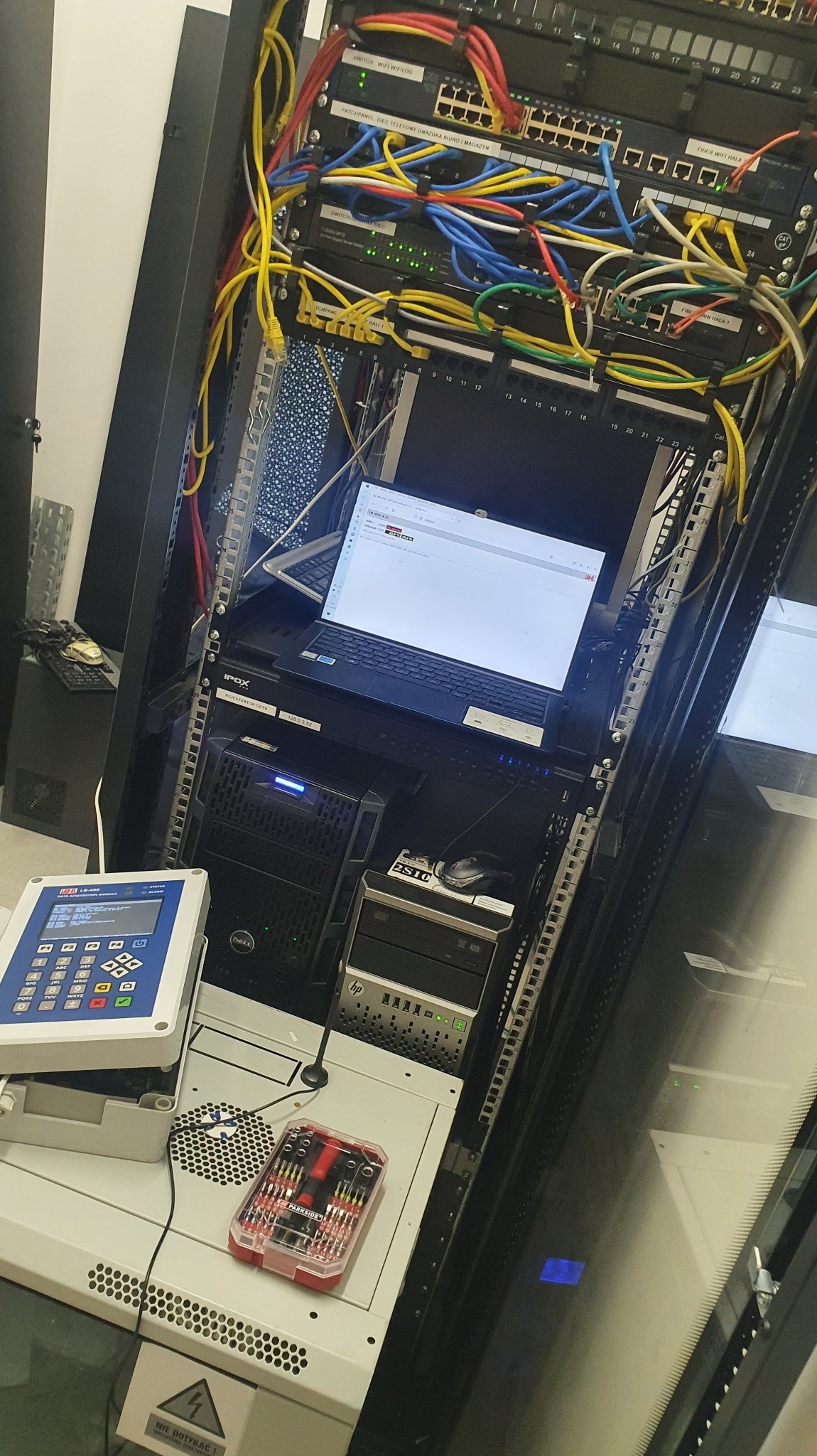 Szafa serwerowa z widocznymi patch panelami, switchami, serwerami Dell i HP, laptopem z uruchomionym interfejsem sieciowym oraz miernikiem sieciowym LB-490 i zestawem narzędzi Parkside.