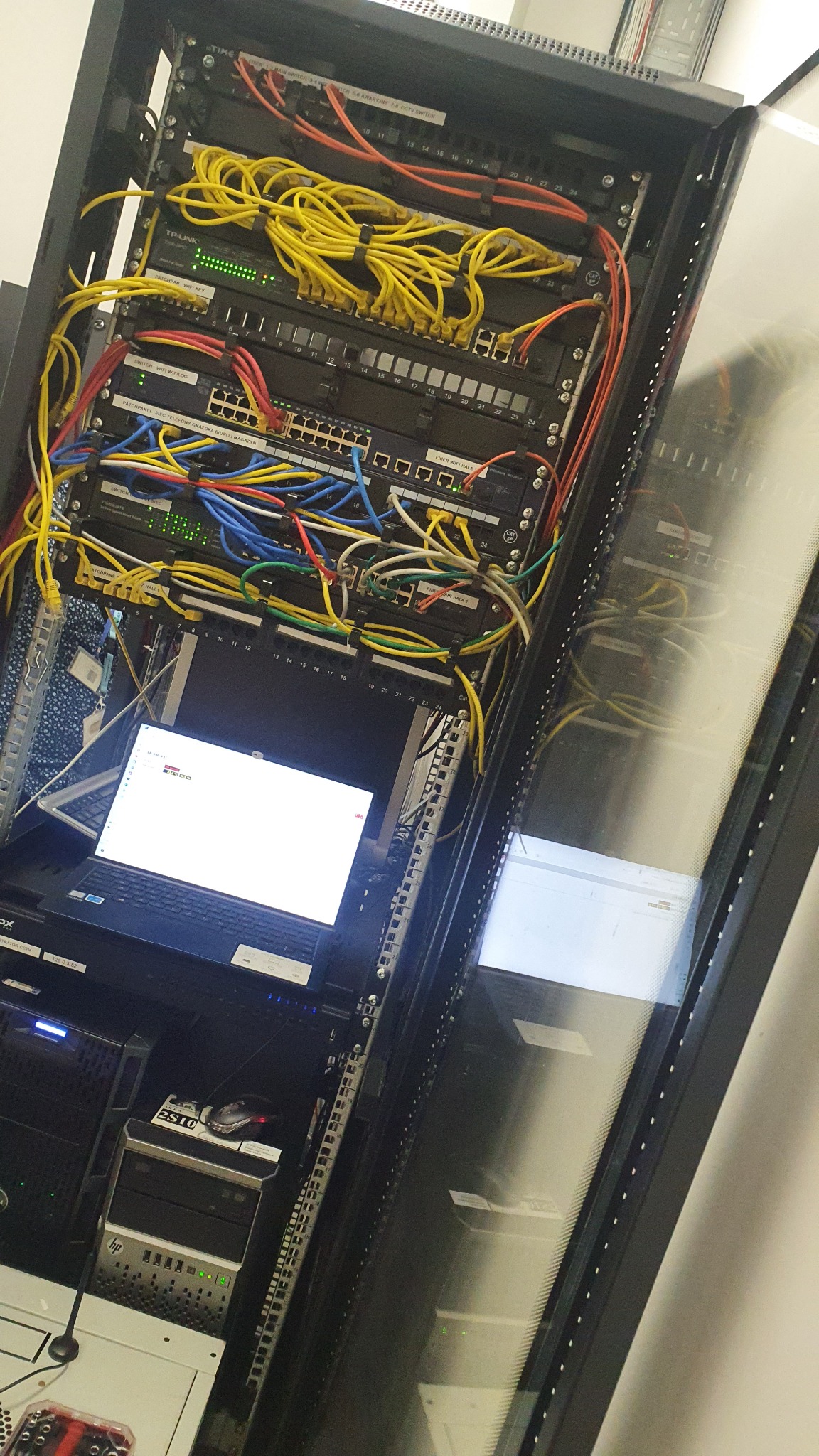 Serwerownia z okablowaniem strukturalnym, switche, patch panele, laptop z włączonym ekranem diagnostycznym, widok od frontu szafy rack z przeszklonymi drzwiami.