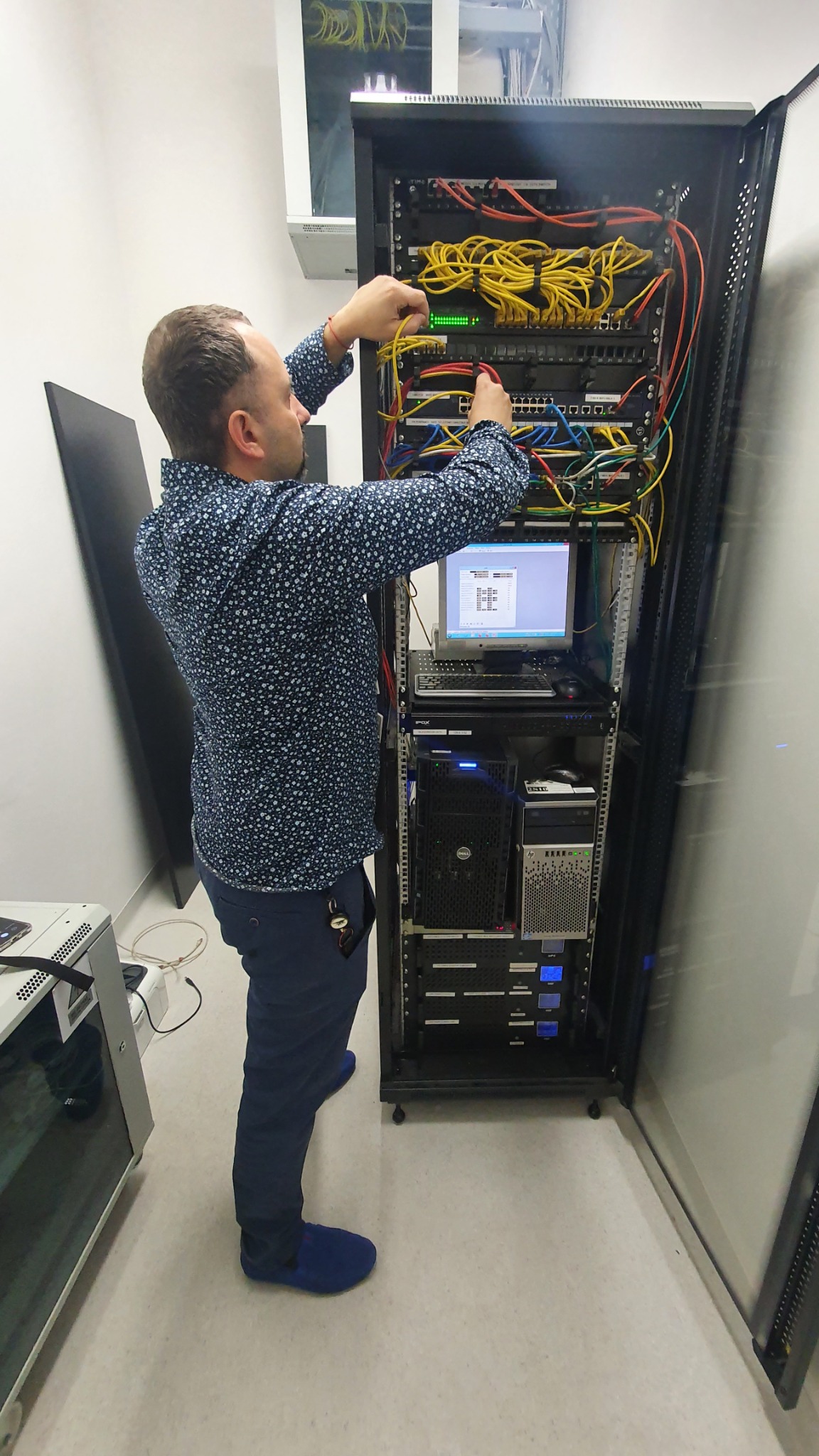 Mężczyzna w koszuli w drobne kwiatki podłącza kable do panelu krosowniczego w szafie serwerowej, obok widoczny monitor i serwery Dell oraz HP.