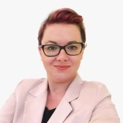 Portret kobiety w okularach, z krótkimi, rudymi włosami, ubranej w jasnoróżową marynarkę, na białym tle.