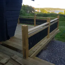 Drewniany taras z czarnymi dekoracyjnymi panelami w balustradzie, widok na morze w tle.