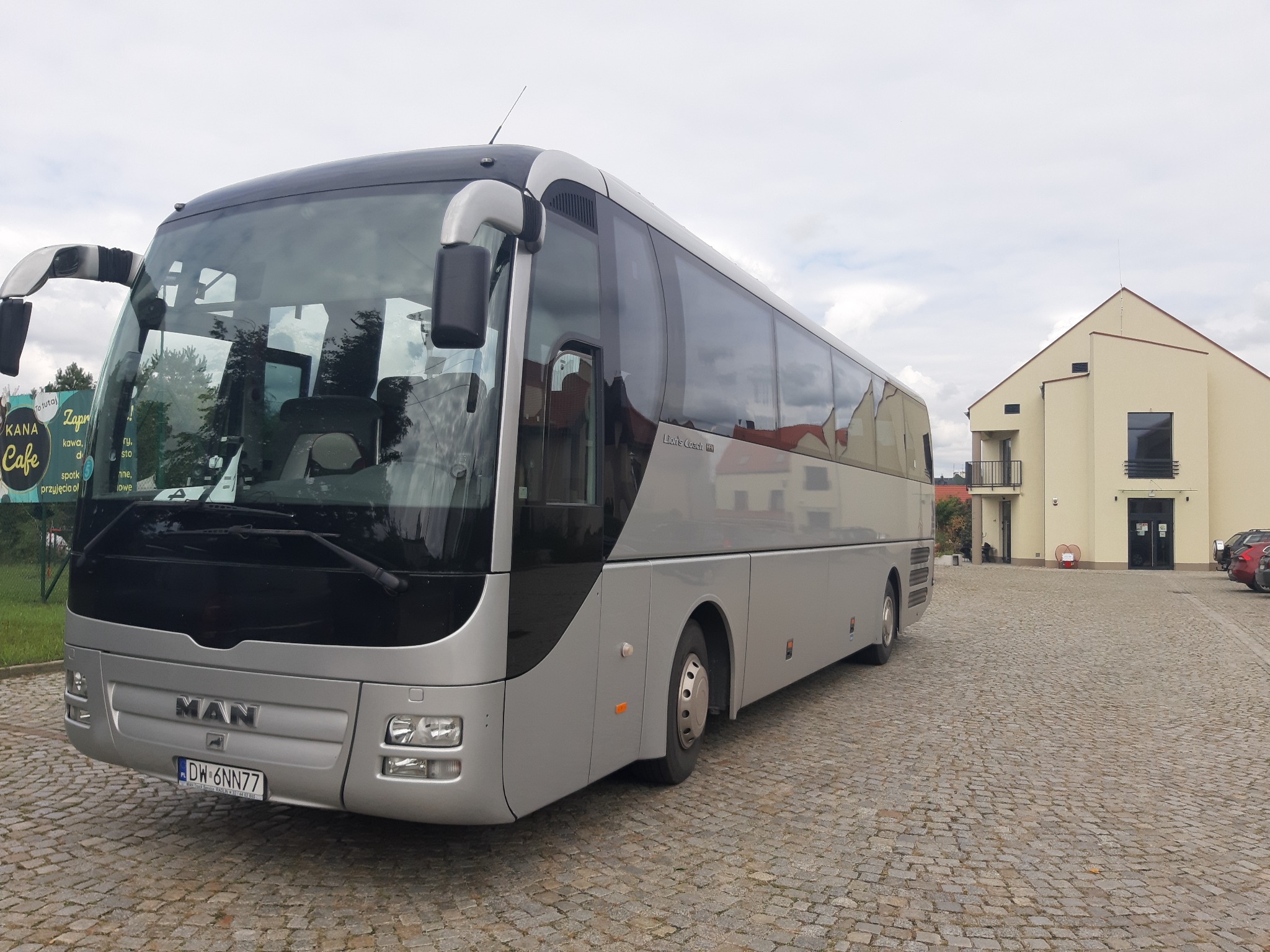 Szary autokar marki MAN Lion's Coach zaparkowany na brukowanym placu, z widoczną tablicą rejestracyjną DW 6NN77 i budynkiem w tle.