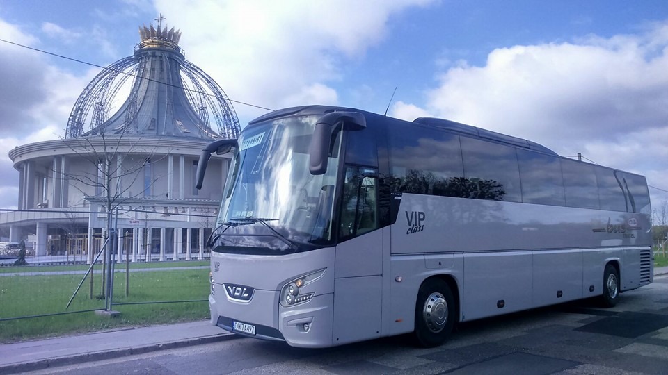 Szary autokar VIP Class marki VDL na tle Bazyliki Najświętszej Maryi Panny Licheńskiej w Licheniu Starym, Polska. Tablica rejestracyjna widoczna.