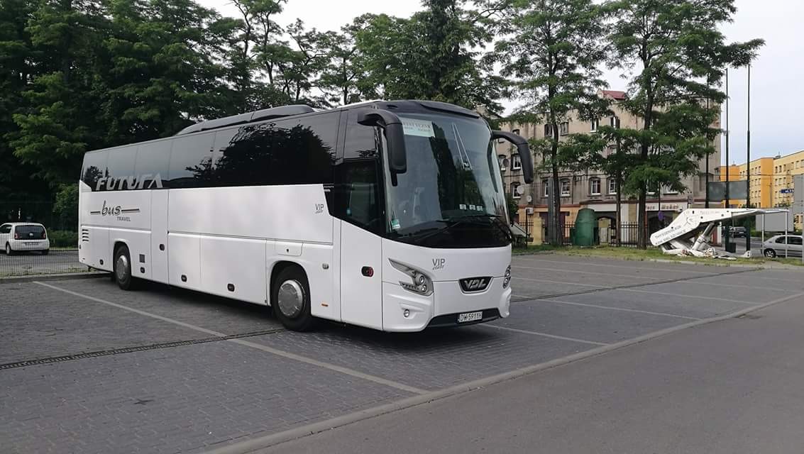 Biały autokar turystyczny marki Futura Bus z czarnymi szybami i oznaczeniem VIP parkuje na pustym parkingu z kostki brukowej, w tle budynki i drzewa.