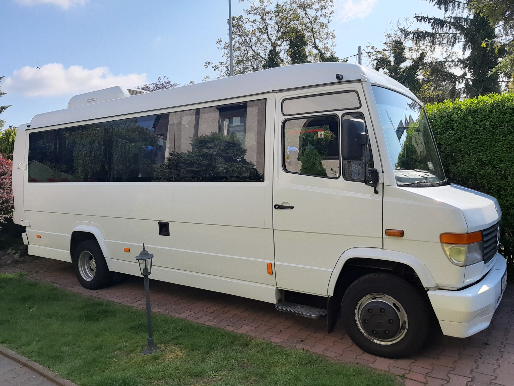 Biały minibus zaparkowany na brukowanym podjeździe, z widoczną lampą ogrodową i bujną zielenią w tle.