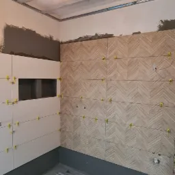 Łazienka w trakcie układania płytek: wzór jodełki na ścianie, białe płytki z wnęką, widoczne krzyżyki dystansowe, surowy beton na podłodze pokryty hydroizolacją, instalacja elektryczna pod sufitem.