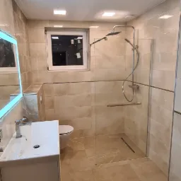 Jasna łazienka z beżowymi płytkami podłogowymi i ściennymi, kabiną prysznicową typu walk-in z odpływem liniowym, umywalką z szafką i podświetlanym lustrem LED oraz oknem z widocznymi naklejkami.