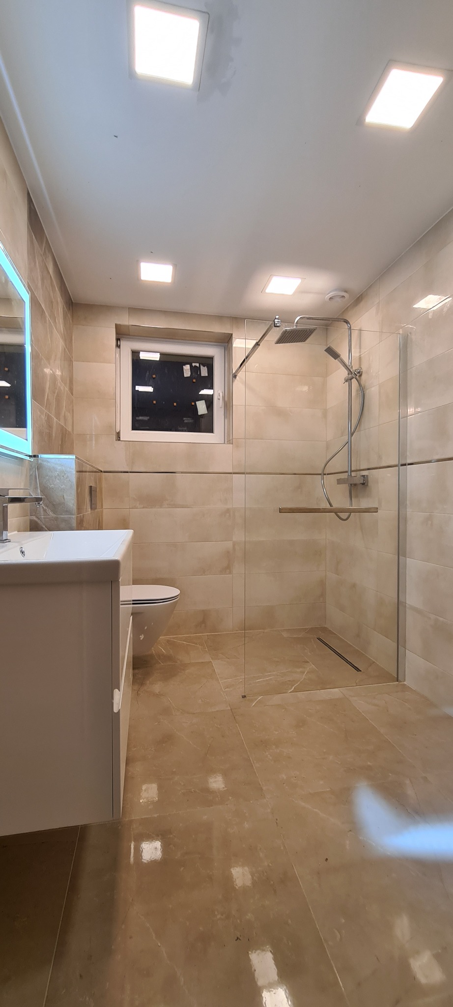 Nowoczesna łazienka z beżowymi płytkami, kabiną prysznicową typu walk-in z deszczownicą, podwieszaną toaletą i białą szafką z umywalką, oświetlona kwadratowymi lampami sufitowymi.