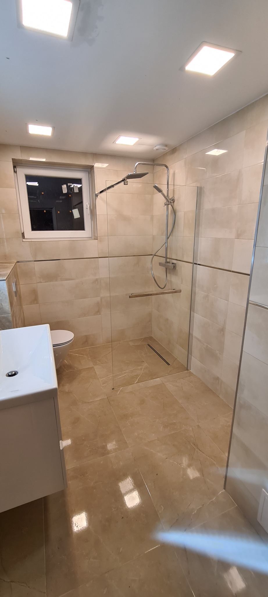 Beżowa łazienka z kabiną prysznicową typu walk-in, umywalką z szafką i wiszącą toaletą, widoczny odpływ liniowy w podłodze.