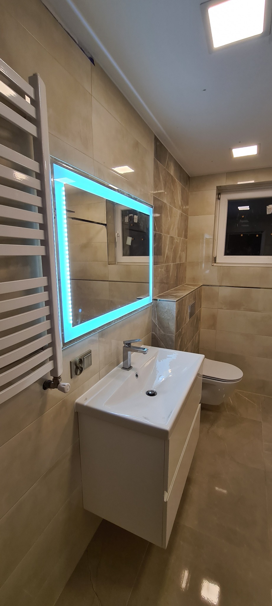 Nowoczesna łazienka z beżowymi płytkami, podwieszaną szafką z umywalką, podświetlanym lustrem LED i grzejnikiem drabinkowym.