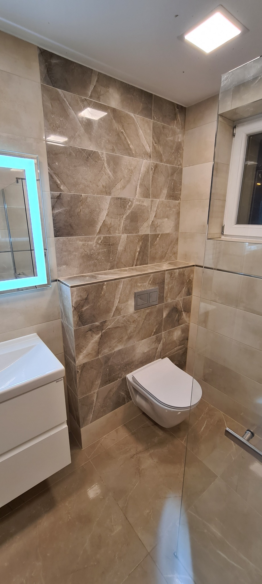 Nowoczesna łazienka w beżowych płytkach imitujących marmur, z podwieszaną toaletą, umywalką z szafką i podświetlanym lustrem.