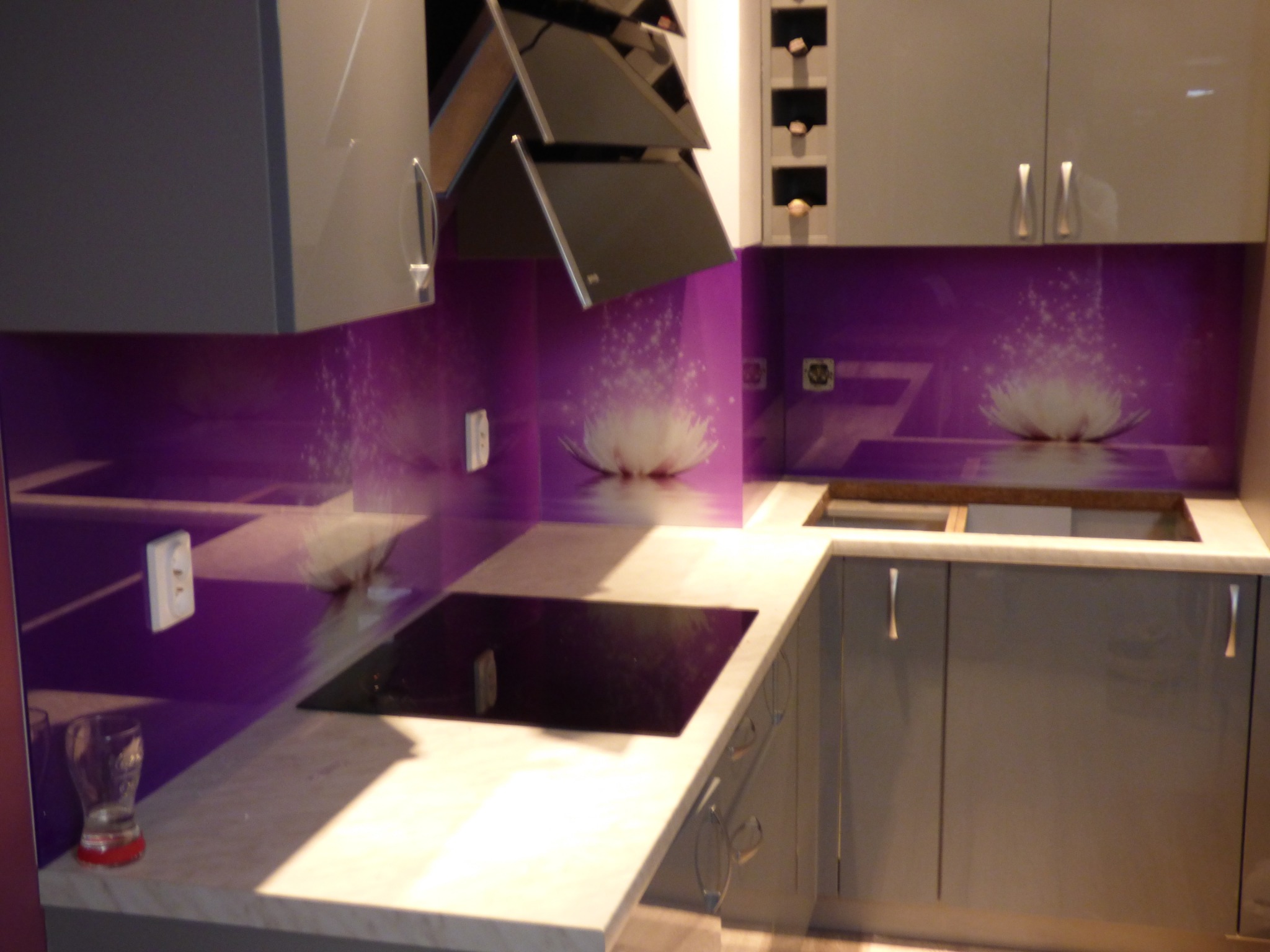 Szklany panel ścienny w kuchni z motywem kwiatu lotosu w fioletowej tonacji, widoczne szafki kuchenne w szarym odcieniu i płyta indukcyjna.