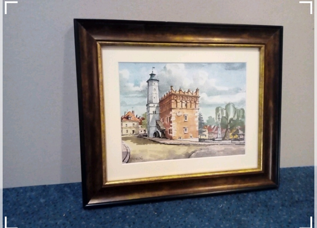 Oprawiony w brązową ramę akwarelowy obraz przedstawiający architekturę miejską z wieżą i budynkiem z blankami, podpisany w lewym dolnym rogu.