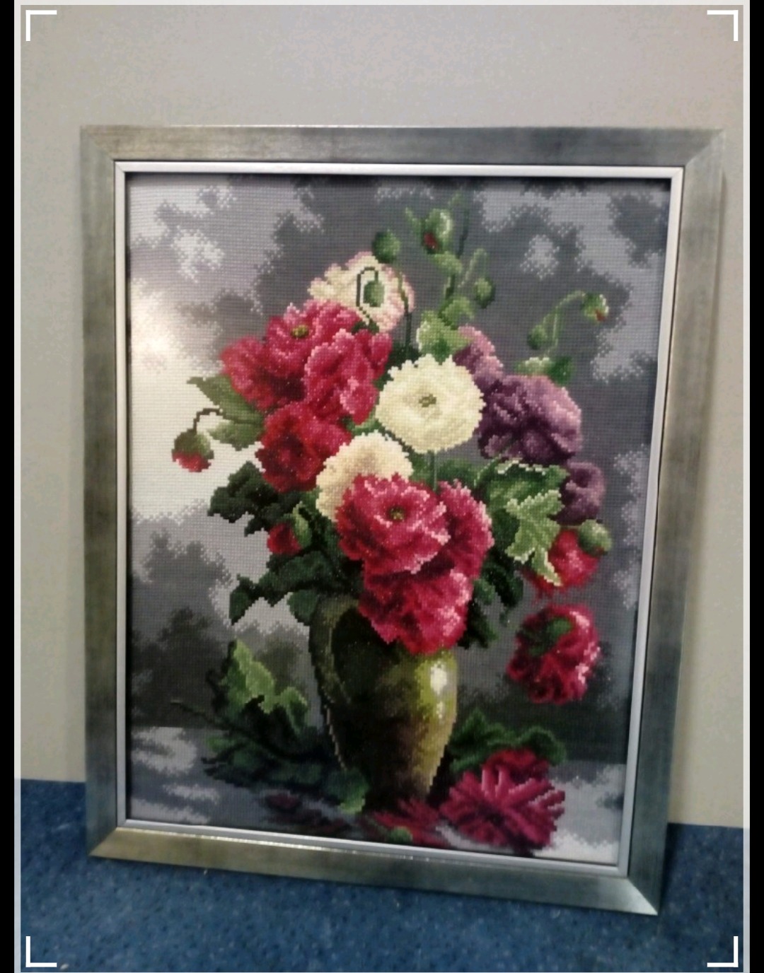Haftowany obraz przedstawiający bukiet kwiatów w wazonie, oprawiony w srebrną ramę, widoczny efekt pikselizacji ściegów.