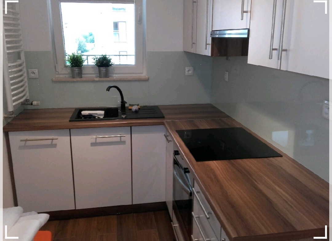 Nowoczesna kuchnia z białymi szafkami, drewnianym blatem i szklanym panelem ściennym w odcieniach szarości, z widocznym oknem z dwoma doniczkami z roślinami.