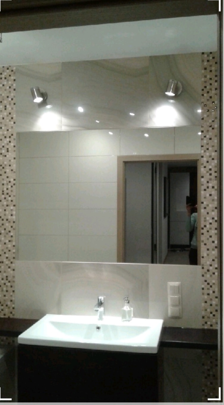 Duże lustro w łazience z mozaikową ramą, odbijające płytki ścienne, oświetlenie punktowe i fragment otwartych drzwi, nad białą umywalką z ciemną szafką i dozownikiem mydła.
