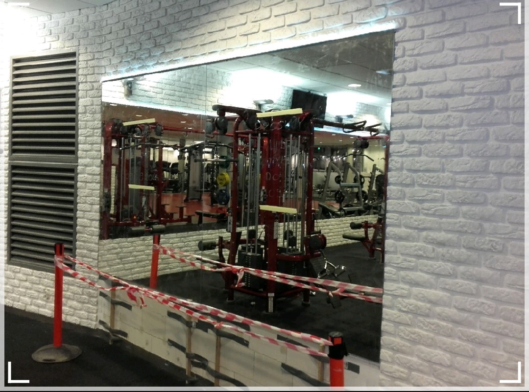 Duże lustro na ścianie z białej cegły w siłowni, odbijające sprzęt do ćwiczeń, z taśmą ostrzegawczą na pierwszym planie.