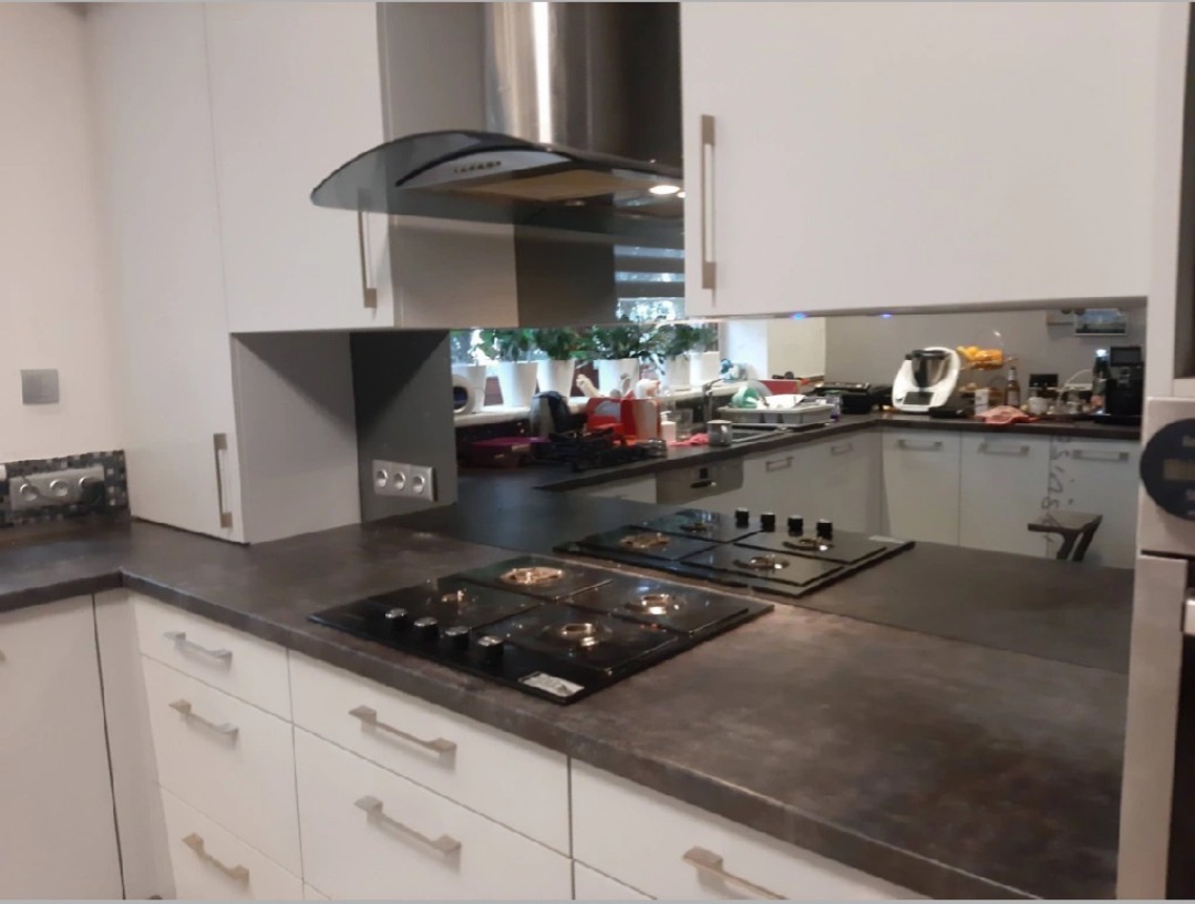 Kuchnia z szarym blatem i białymi szafkami; lustrzane panele między szafkami górnymi a blatem, odbijające fragment wnętrza z roślinami w doniczkach.