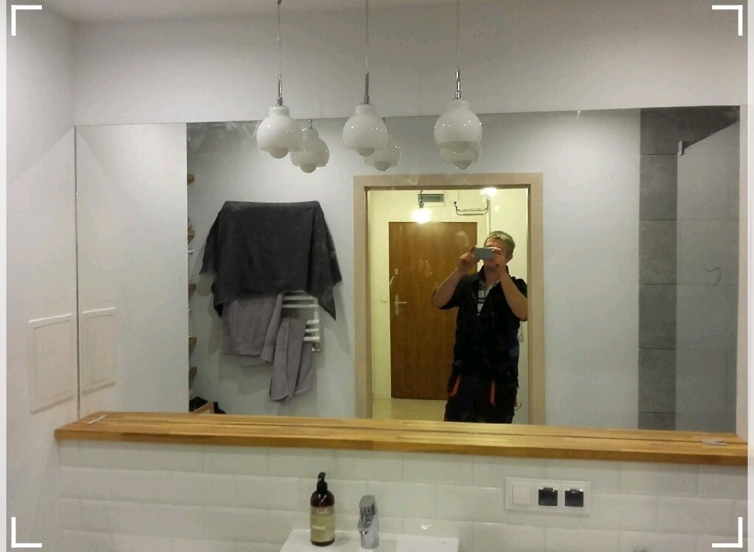 Duże lustro łazienkowe zamontowane nad umywalką z drewnianym blatem, widoczne wiszące lampy i odbicie osoby robiącej zdjęcie telefonem.