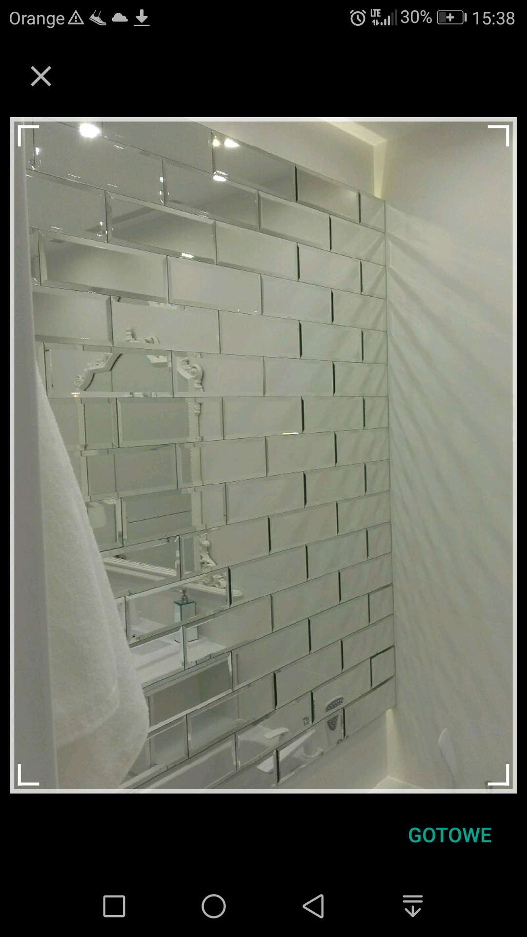 Ściana w łazience wyłożona prostokątnymi lustrami, z widocznym fragmentem wanny i dozownikiem mydła odbitym w lustrach, biała ściana po prawej stronie.