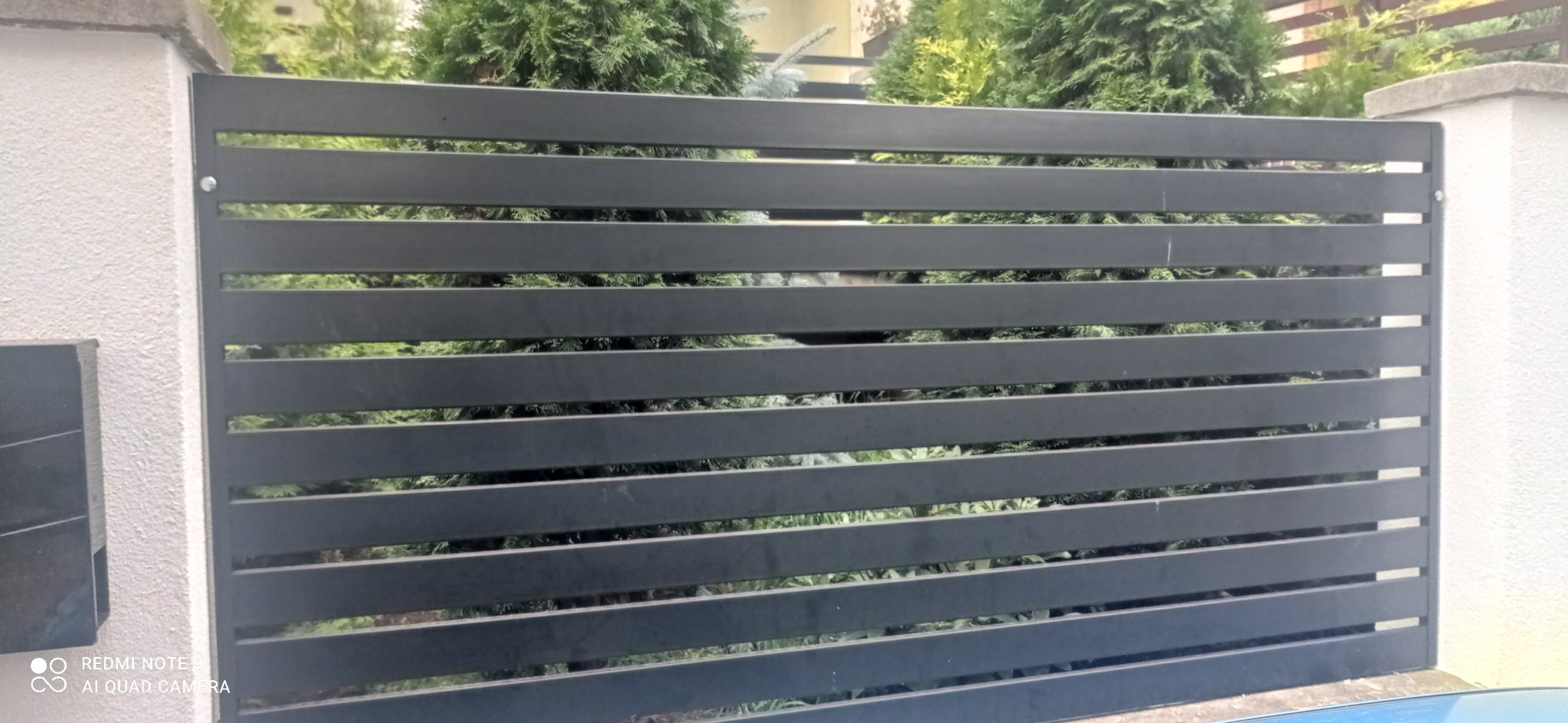 Nowoczesne, poziome przęsło ogrodzeniowe w kolorze grafitowym, zamontowane między białymi słupkami, z widoczną zielenią roślin w tle.