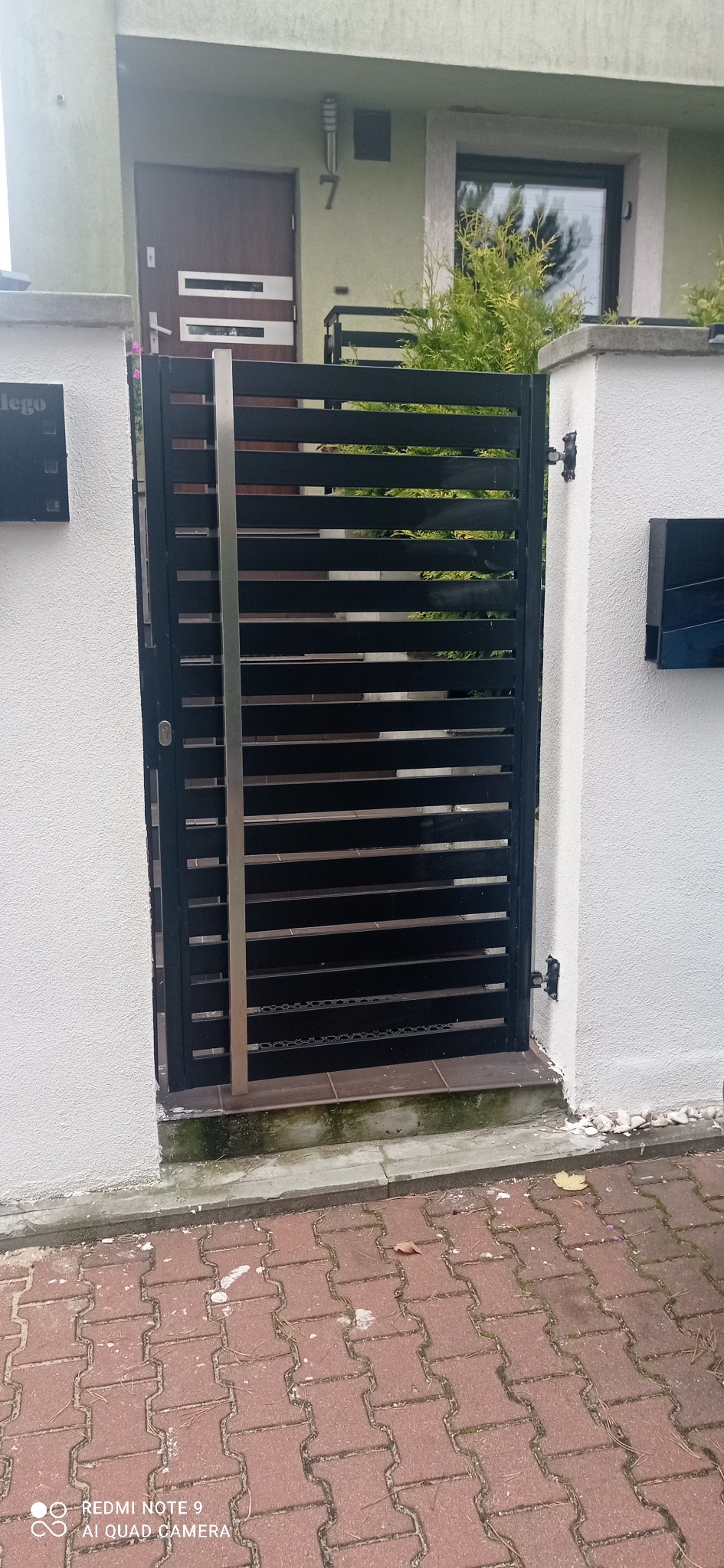 Nowoczesna furtka z poziomych, czarnych profili metalowych z pionowym akcentem ze stali nierdzewnej, zamontowana między dwoma białymi słupkami ogrodzeniowymi, z widoczną kostką brukową i fragmentem...