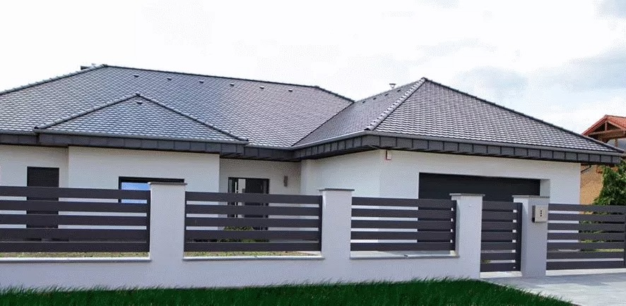 Nowoczesne ogrodzenie posesji z poziomych, grafitowych paneli między białymi słupkami, na tle domu z ciemną dachówką.
