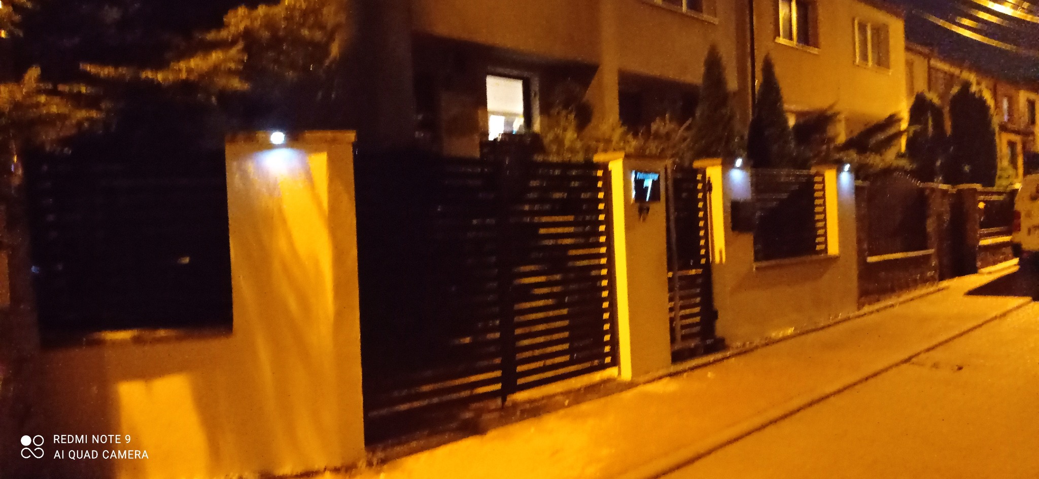 Nowoczesne ogrodzenie z poziomych, czarnych paneli metalowych osadzonych w jasnych, oświetlonych słupkach, widziane nocą z poziomu ulicy przed domem.