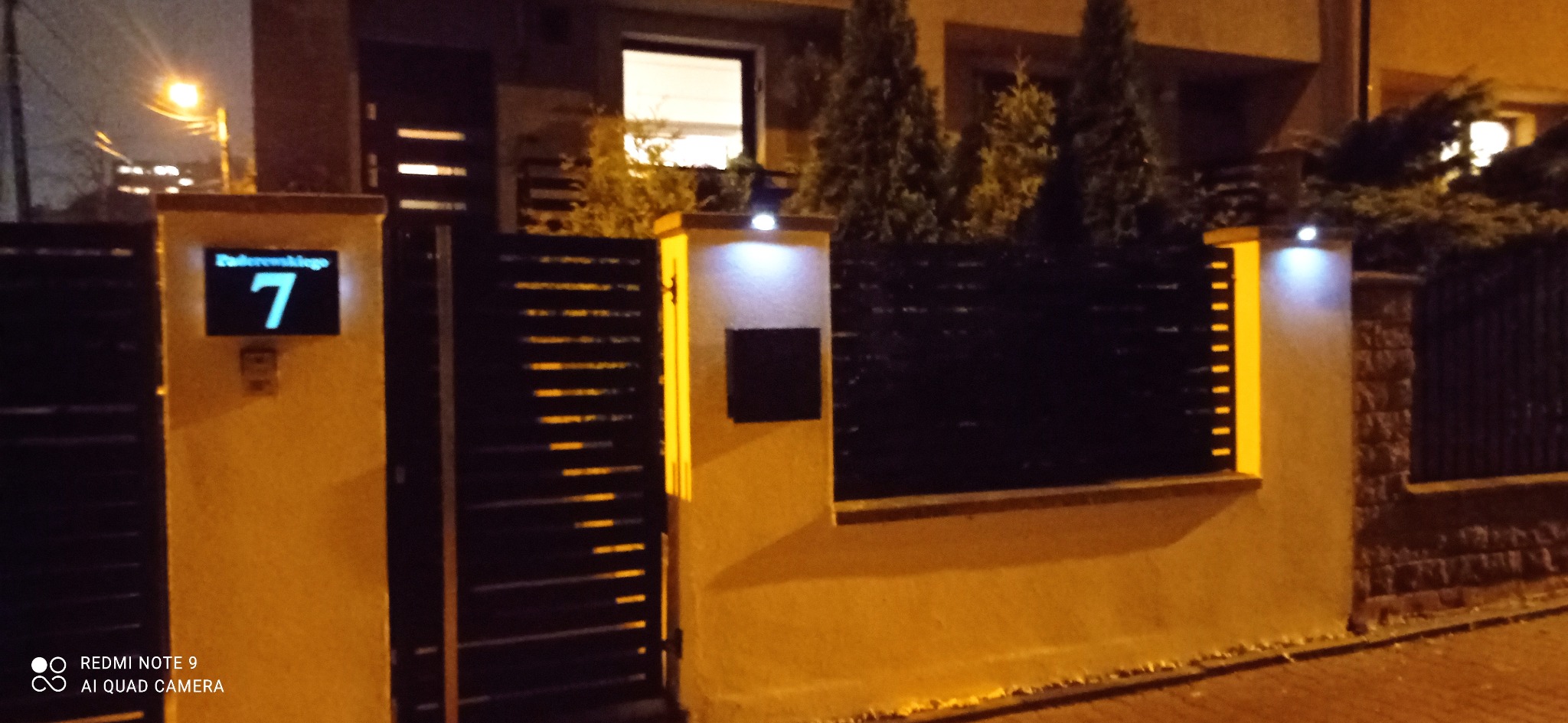 Nowoczesne ogrodzenie z poziomych metalowych paneli, oświetlone lampkami LED, z numerem domu podświetlonym na słupku wejściowym.