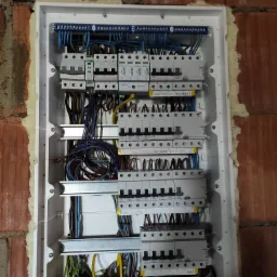 Rozdzielnica elektryczna w trakcie montażu, widoczne liczne przewody, zabezpieczenia i listwy zaciskowe, osadzona w murze z cegły z widoczną pianką montażową.