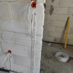 Instalacja elektryczna w trakcie montażu: białe przewody prowadzone wzdłuż ściany z betonu komórkowego, widoczne puszki instalacyjne w kolorze pomarańczowym i poziomica oparta o ścianę w tle.
