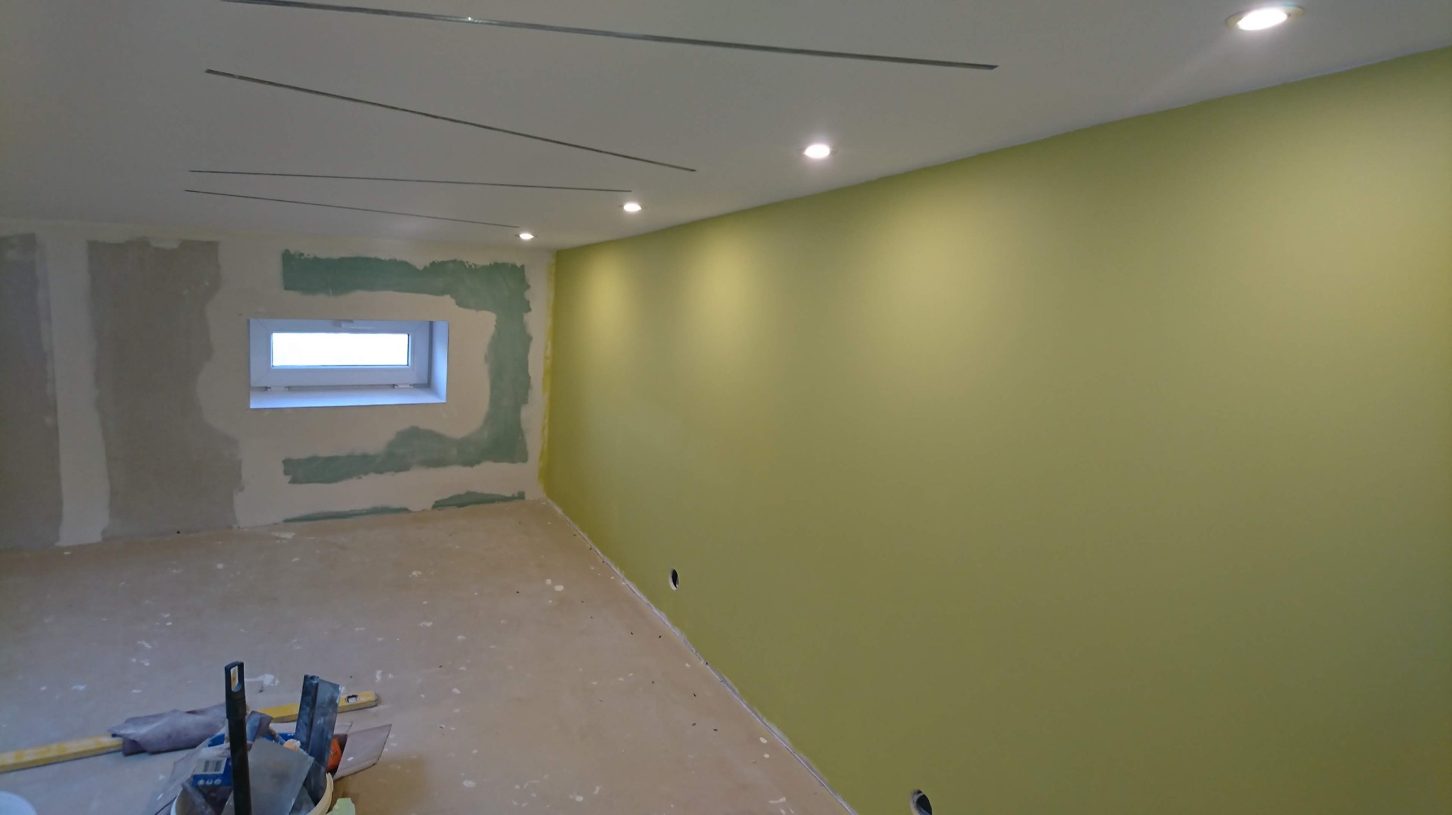 Pokój w trakcie remontu: zielona, pomalowana ściana, surowy fragment ściany z oknem, częściowo wyszpachlowany, metalowe listwy na suficie pod oświetlenie, narzędzia na podłodze.