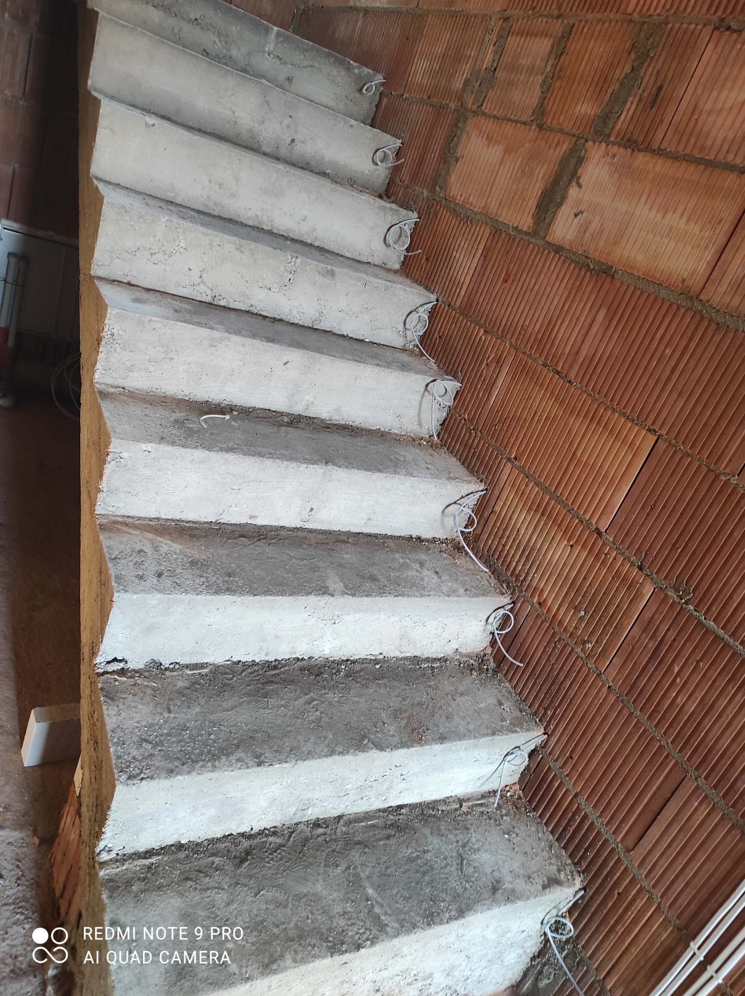 Surowe, betonowe schody wewnętrzne w trakcie budowy, z wystającymi przewodami elektrycznymi przy ścianie z cegły dziurawki.