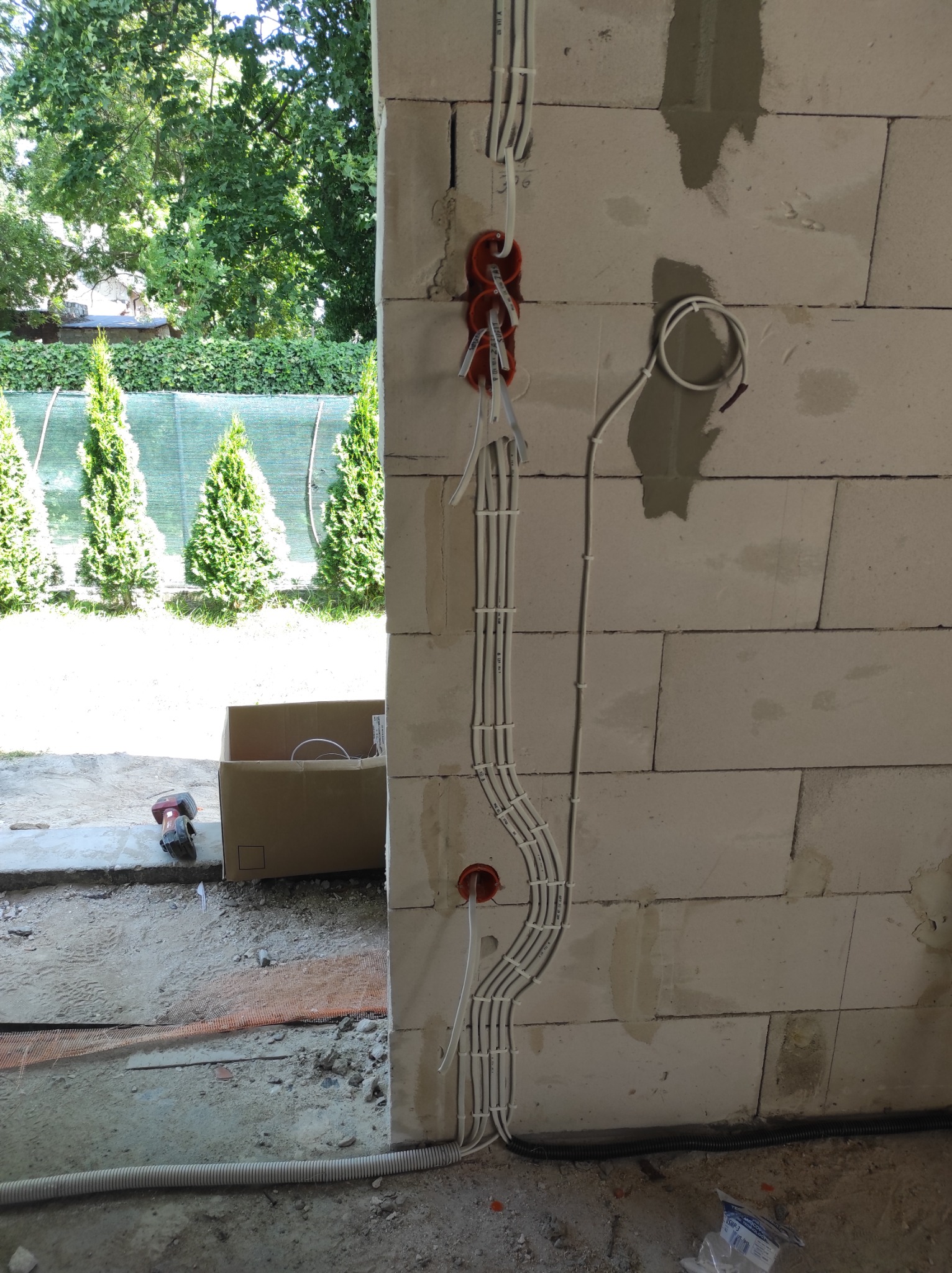 Ściana z betonu komórkowego w trakcie prac instalacyjnych: widoczne kable elektryczne prowadzone w peszlach, puszki instalacyjne w kolorze pomarańczowym, częściowo zamurowane bruzdy, w tle...