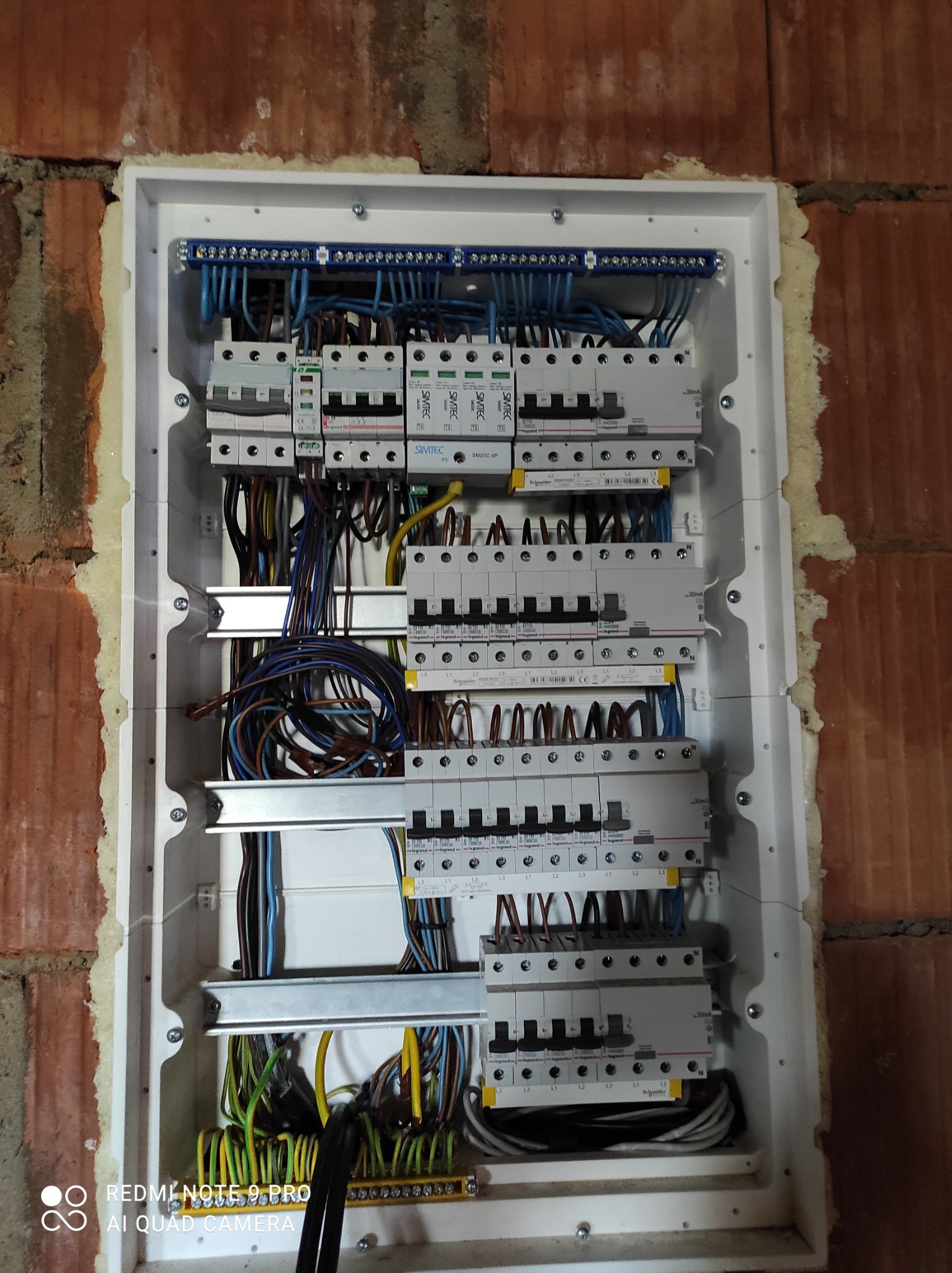 Rozdzielnica elektryczna w trakcie montażu, widoczne liczne przewody, zabezpieczenia i listwy zaciskowe, osadzona w murze z cegły z widoczną pianką montażową.