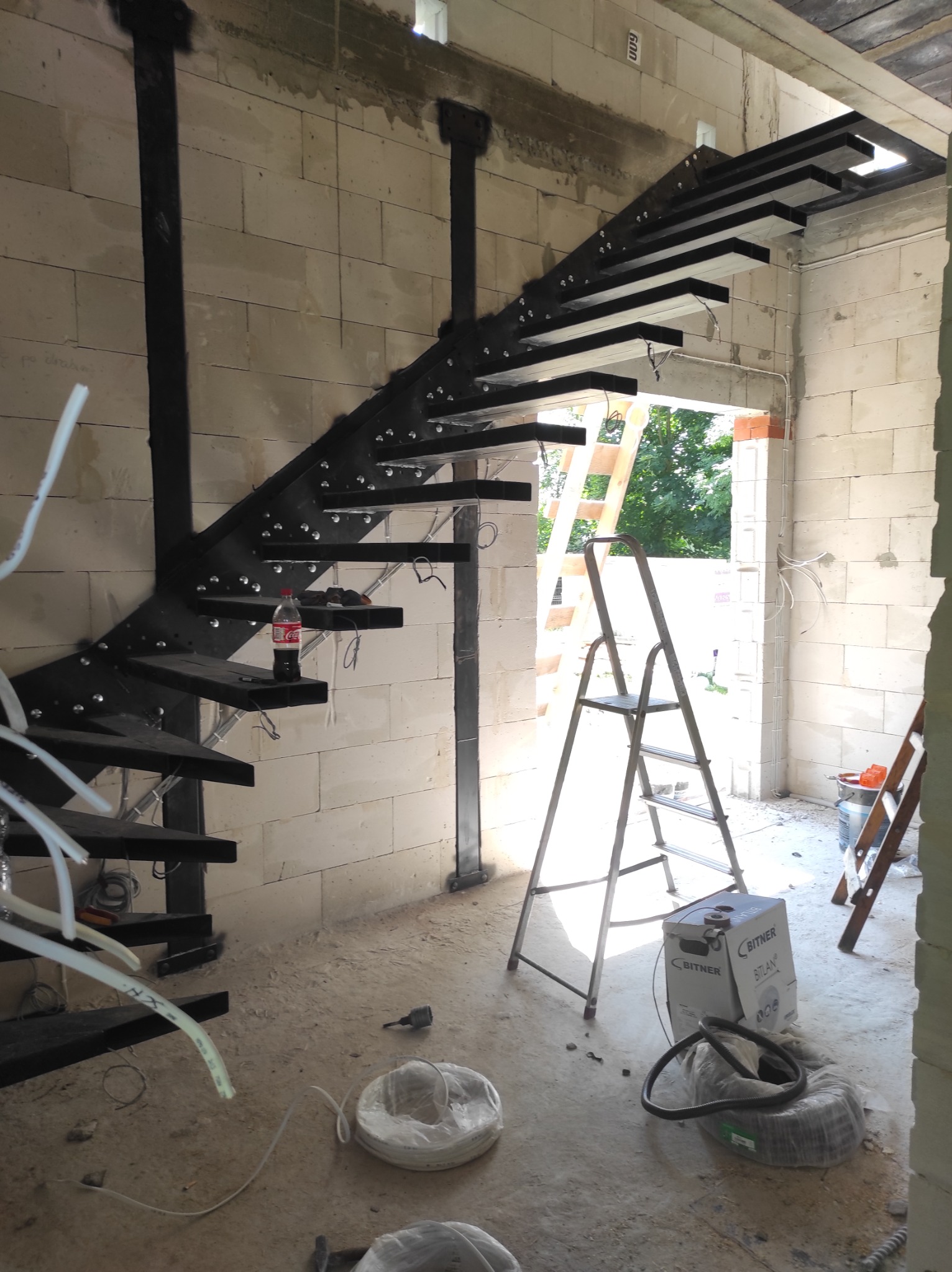 Wnętrze w trakcie budowy z spiralnymi schodami stalowymi, butelką Coca-Coli na jednym ze stopni, widocznymi instalacjami elektrycznymi i drabiną opartą o framugę drzwi.