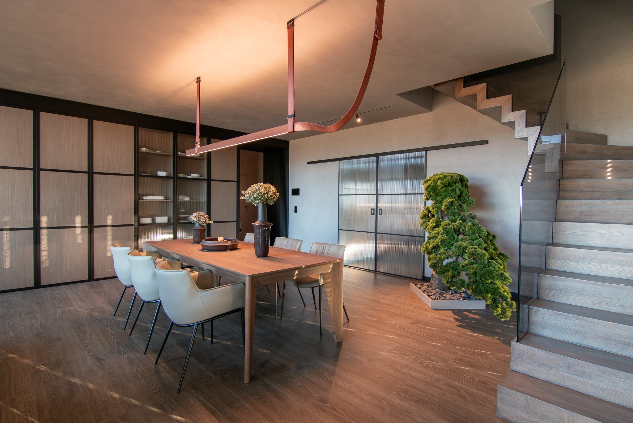 Nowoczesna jadalnia z drewnianym stołem, krzesłami w odcieniach beżu, przeszklonymi drzwiami przesuwnymi i schodami z drewnianymi stopniami.