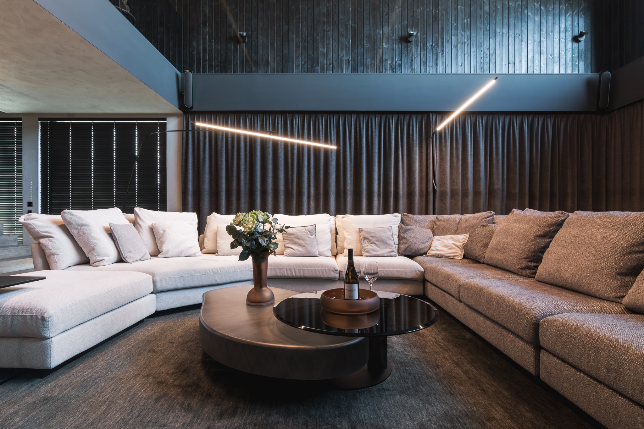 Nowoczesny salon z narożną sofą, stolikiem kawowym z butelką wina i kwiatami. Ciemne drewniane ściany i designerskie oświetlenie tworzą przytulną atmosferę.