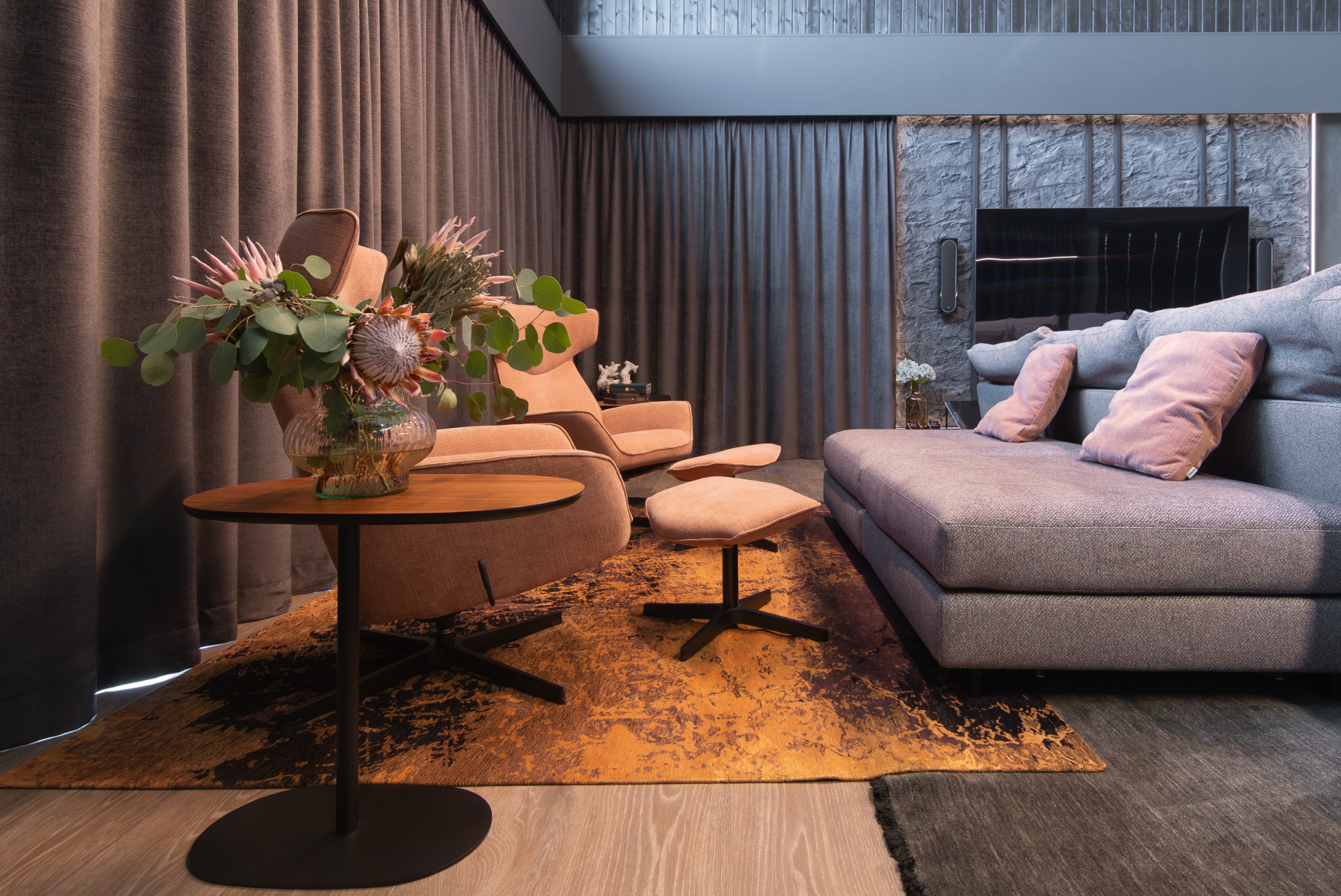 Stylowy salon z fotelami w odcieniach brzoskwini, szarą sofą i stolikiem z kwiatami. Dywan w odcieniach złota i brązu dodaje ciepła. Ściana z kamienia za sofą.