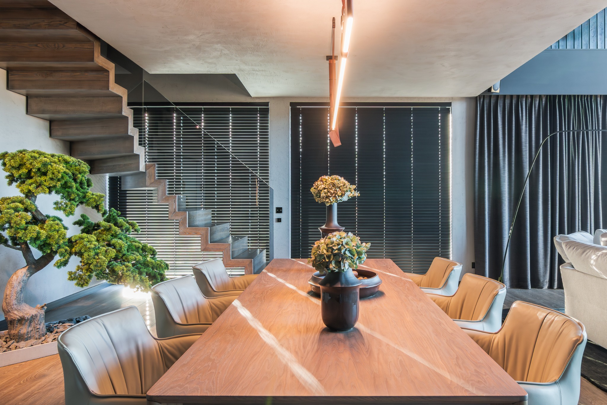 Nowoczesna jadalnia z drewnianym stołem, krzesłami w odcieniach beżu i szarości, schodami oraz bonsai. Minimalistyczne oświetlenie i zasłony w tle.