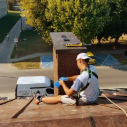 Dekarz w uprzęży siedzi na miedzianym dachu, zakłada niebieskie rękawice, w tle budynki mieszkalne i drzewa, widoczny samochód dostawczy z napisem 'Usługi Budowlane'.