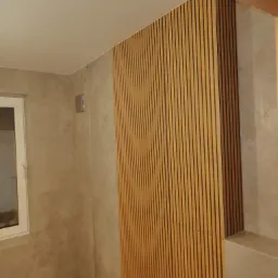 Ściana w łazience wykończona panelami z drewnianych listewek, sąsiadująca z płytkami imitującymi beton i białym oknem.